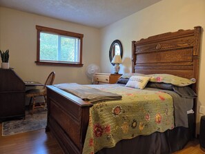 Un dormitorio con una cama de madera, un escritorio con silla, una ventana con persianas, una mesita de noche con lámpara y un espejo en la pared.