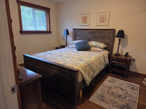 A bedroom with a wooden bed, two bedside lamps, a nightstand, a window with blinds, and two framed pictures on the wall. - Stunning 2BR cottage on Vancouver Island in Natural Adventure Paradise! (Lake Cowichan)