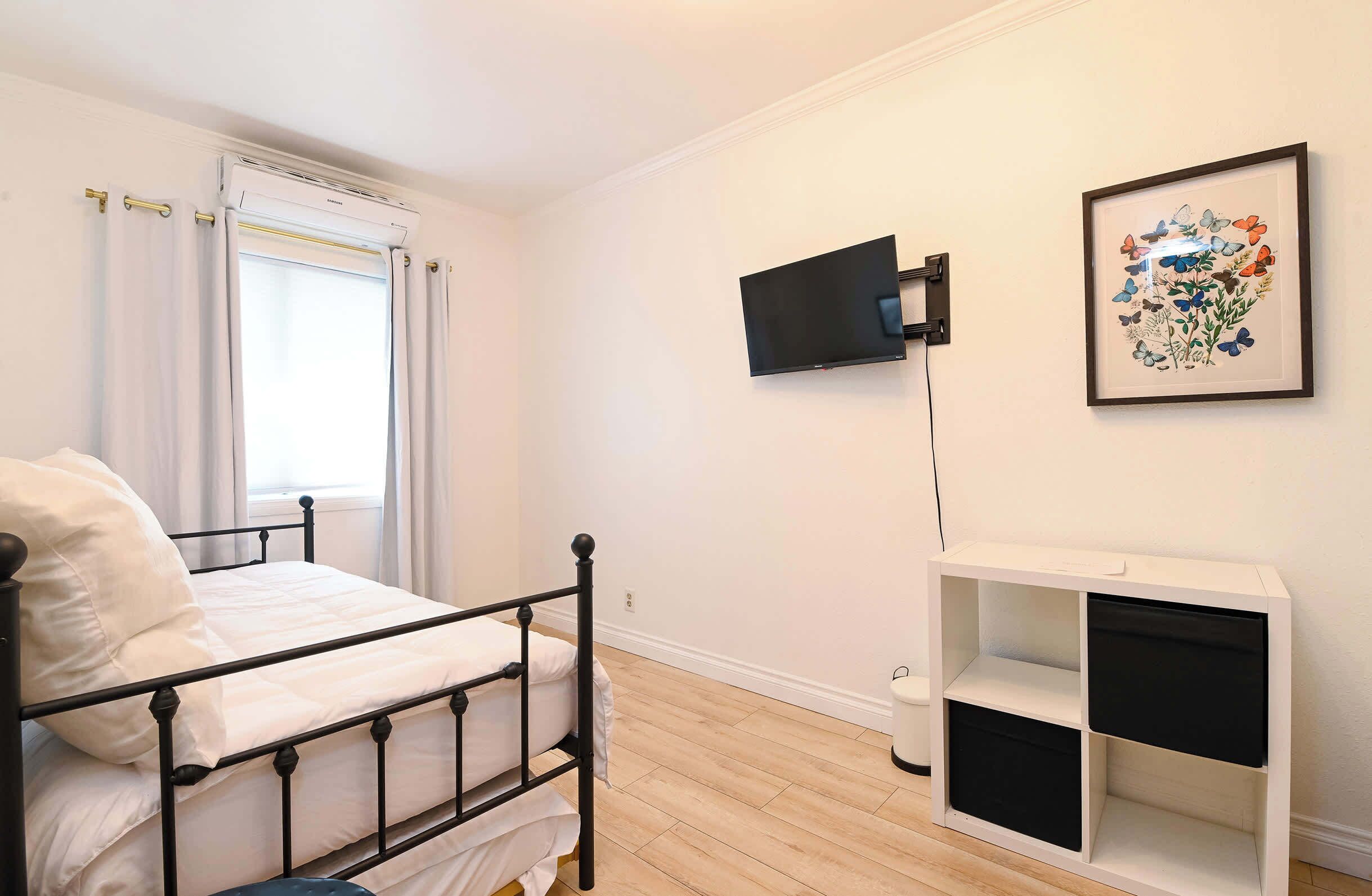 A bedroom with a bed, a TV mounted on the wall, a framed floral artwork, and a white storage unit.