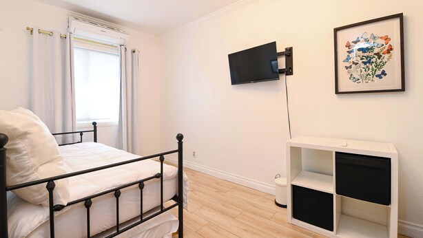 A bedroom with a bed, a TV mounted on the wall, a framed floral artwork, and a white storage unit. - Immaculate 3bdr Private Bungalow, Walk to Beach! (Long Beach)