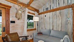 A rustic cabin interior with a mounted deer head, a grey sofa, a brown leather chair, and a wooden ceiling. - The Hendo - Cabin #1 - Studio Style (Henderson)