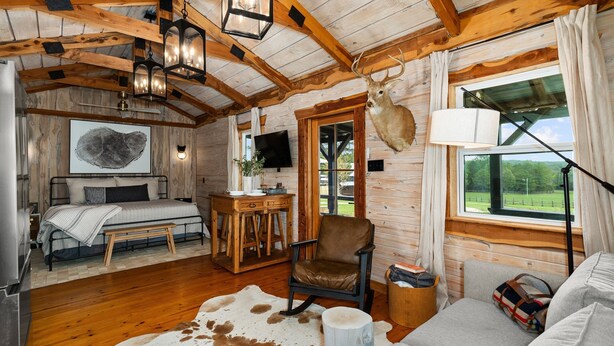 A cozy cabin interior with a bed, a dining area, a mounted deer head, and a large window offering a view of the outdoors. - The Hendo - Cabin #1 - Studio Style (Henderson)