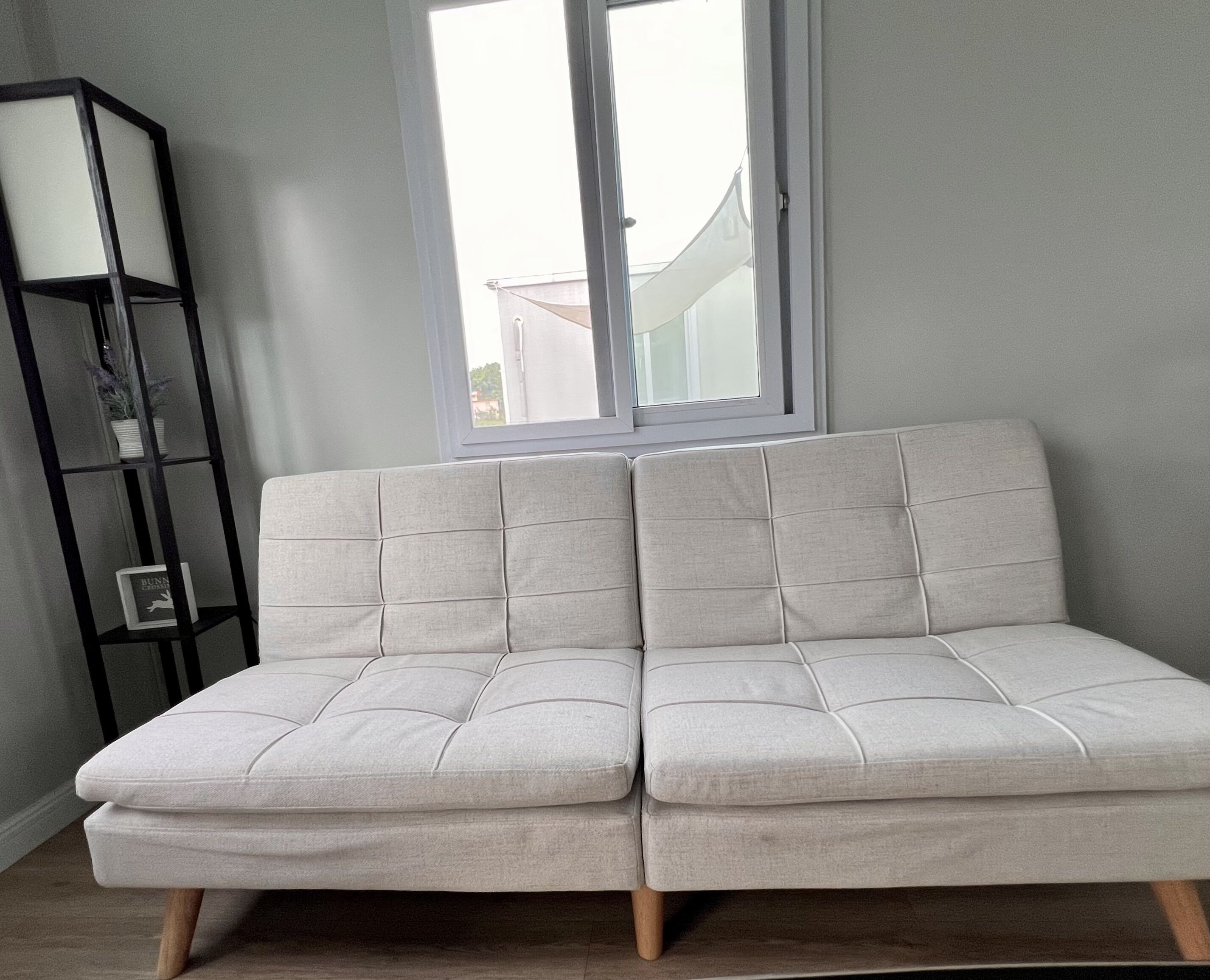 Ein helles Sofa mit Holzbeinen in einem Zimmer mit Fenster und einer schwarzen Regaleinheit.