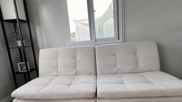 Ein helles Sofa mit Holzbeinen in einem Zimmer mit Fenster und einer schwarzen Regaleinheit.