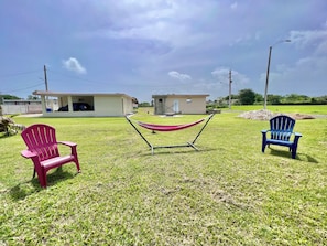 Un espacio al aire libre con césped, dos sillas Adirondack, una hamaca y un edificio pequeño al fondo.