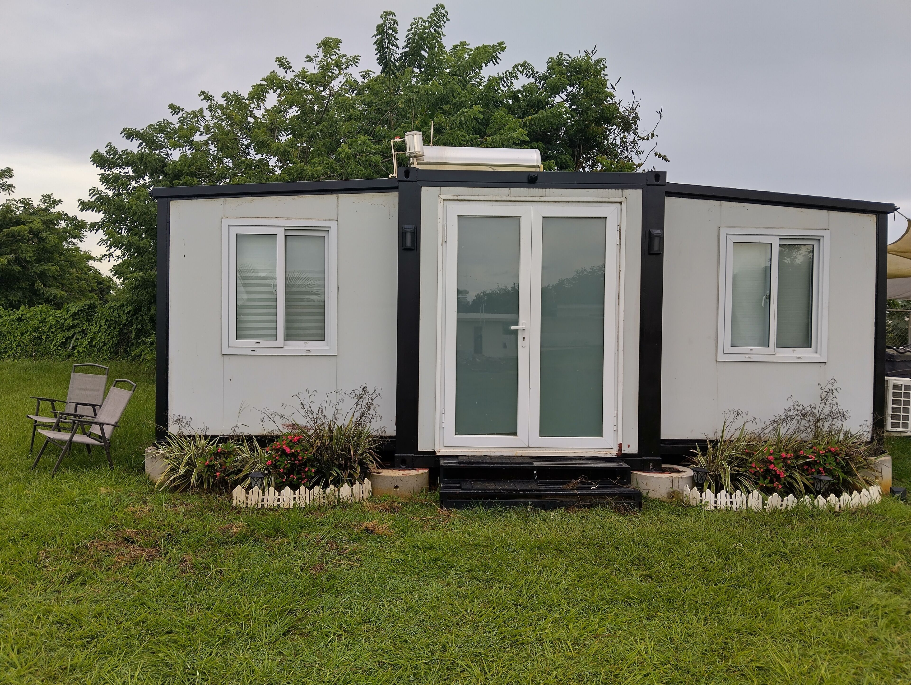 Une petite maison de plain-pied, à l’extérieur blanc, dotée d’une porte-fenêtre coulissante et d’un petit porche avec une chaise et des plantes en pot.