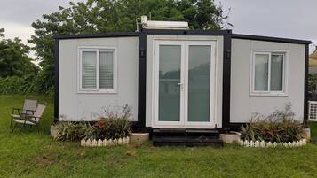 Une petite maison de plain-pied, à l’extérieur blanc, dotée d’une porte-fenêtre coulissante et d’un petit porche avec une chaise et des plantes en pot.