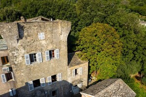 A multi-story building with a rustic stone facade, multiple windows with shutters, and a surrounding dense forest.