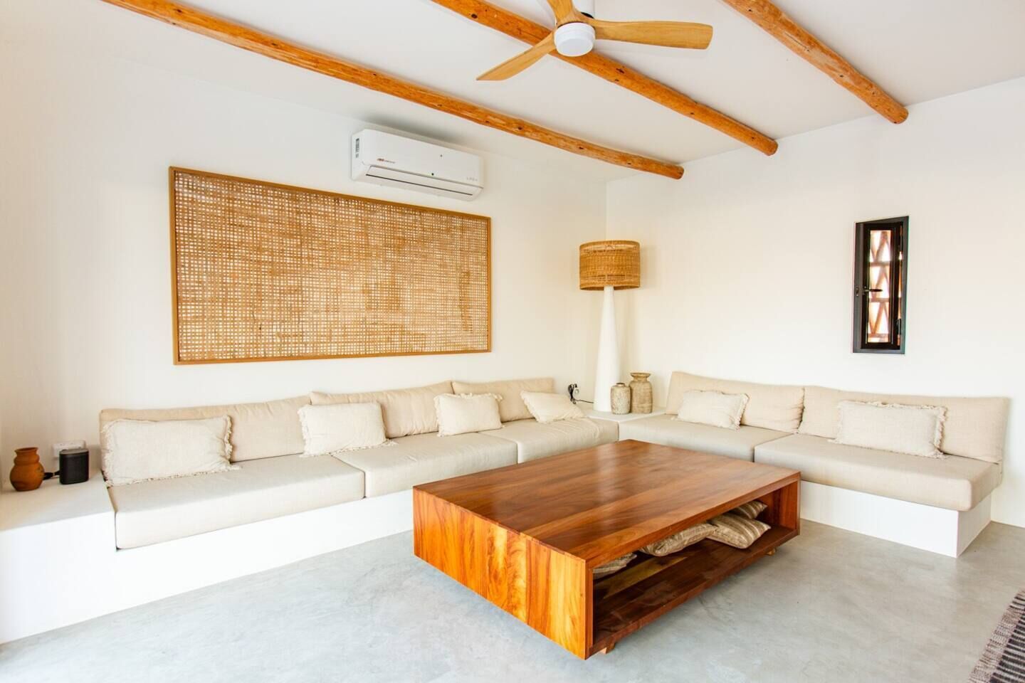 Un salon comprenant une table basse en bois, un ensemble de canapés, une décoration murale en fibres tressées, une lampe sur pied et un appareil de climatisation.
