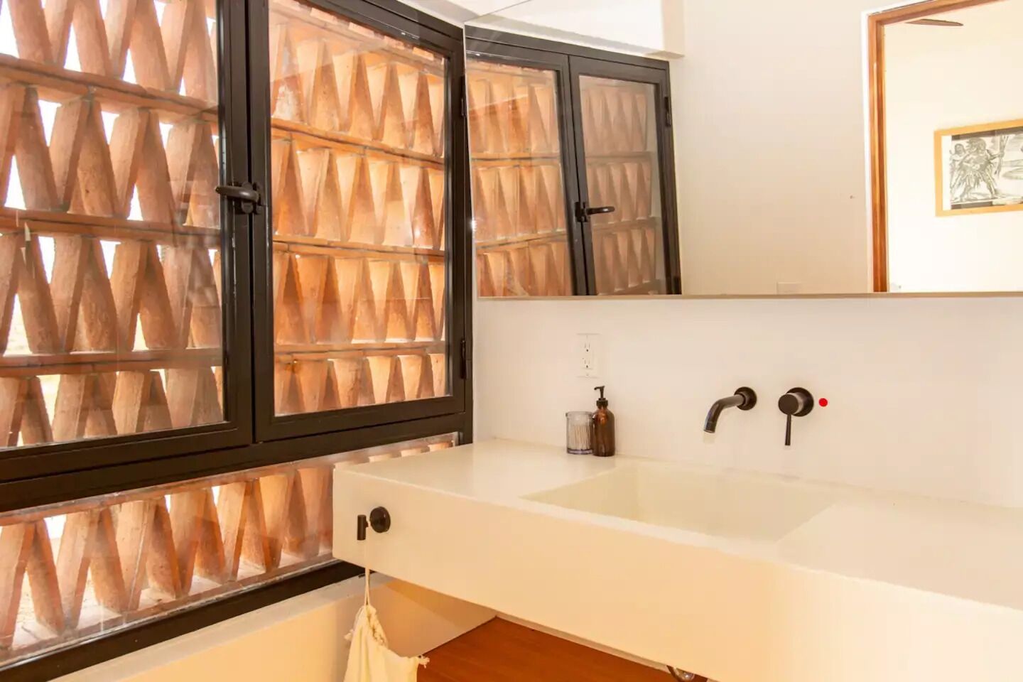 A bathroom with a white sink, two faucets, a mirror, and a window with a geometric patterned blind.