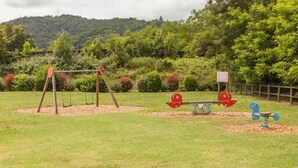 Un parque infantil con columpios, un balancín y una estructura para escalar, en un área verde con árboles y una colina al fondo.