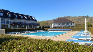 Zona de piscina con sillones de descanso y sombrillas, rodeada de setos cuidados y edificios con balcones.