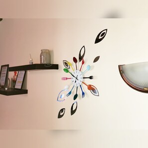 Un reloj de pared con un diseño original que utiliza utensilios de cocina como indicadores de las horas, un estante con un tarro y una botella, y una lámpara.