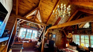 L’intérieur d’un chalet confortable, avec des murs en rondins, un plafond haut, un lustre, un lit recouvert d’une couverture à motifs, un fauteuil à bascule et un chat perché sur une poutre.