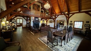 Une pièce spacieuse avec un plafond haut et des poutres apparentes, comprenant une cheminée, une table à manger avec des chaises et un lustre.