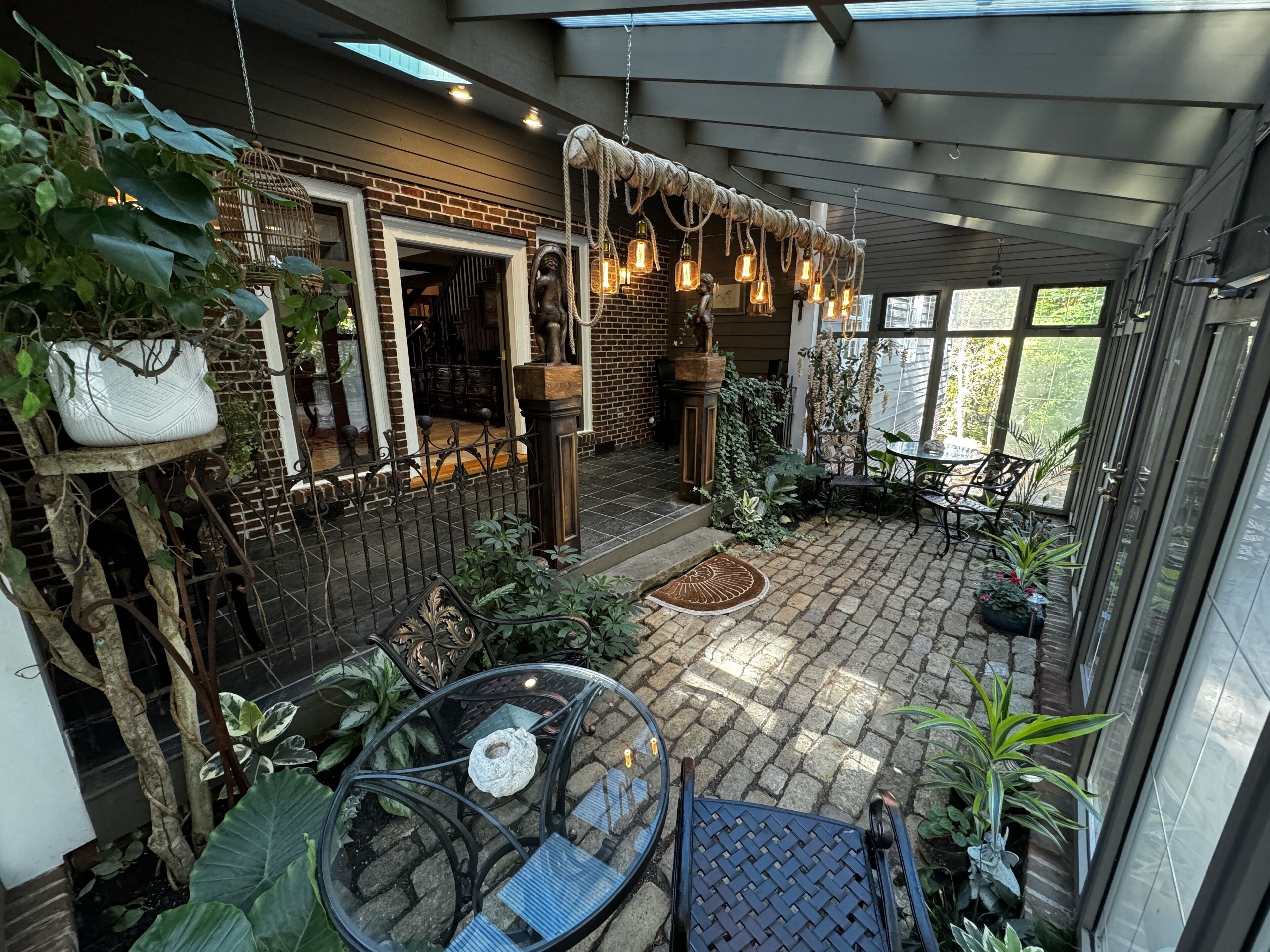 A covered patio area with brick flooring, a glass table, and various potted plants.