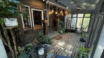 A covered patio area with brick flooring, a glass table, and various potted plants.
