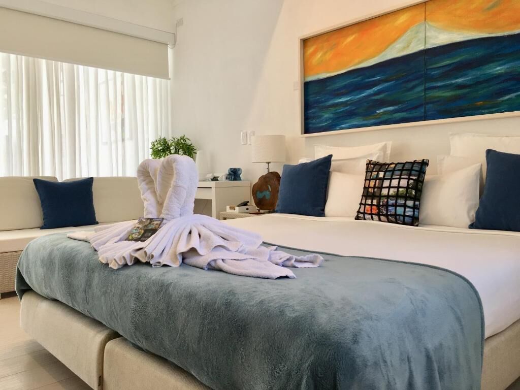 A hotel room with a large bed, a painting of a sunset over the ocean, and a window with sheer curtains.