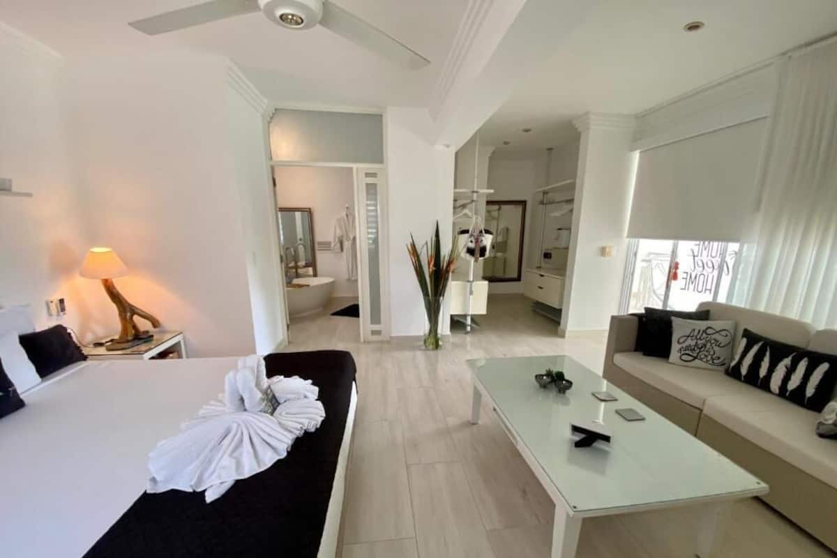 A modern hotel room with a bed, a sofa, a glass coffee table, and a view of the hallway and other rooms.