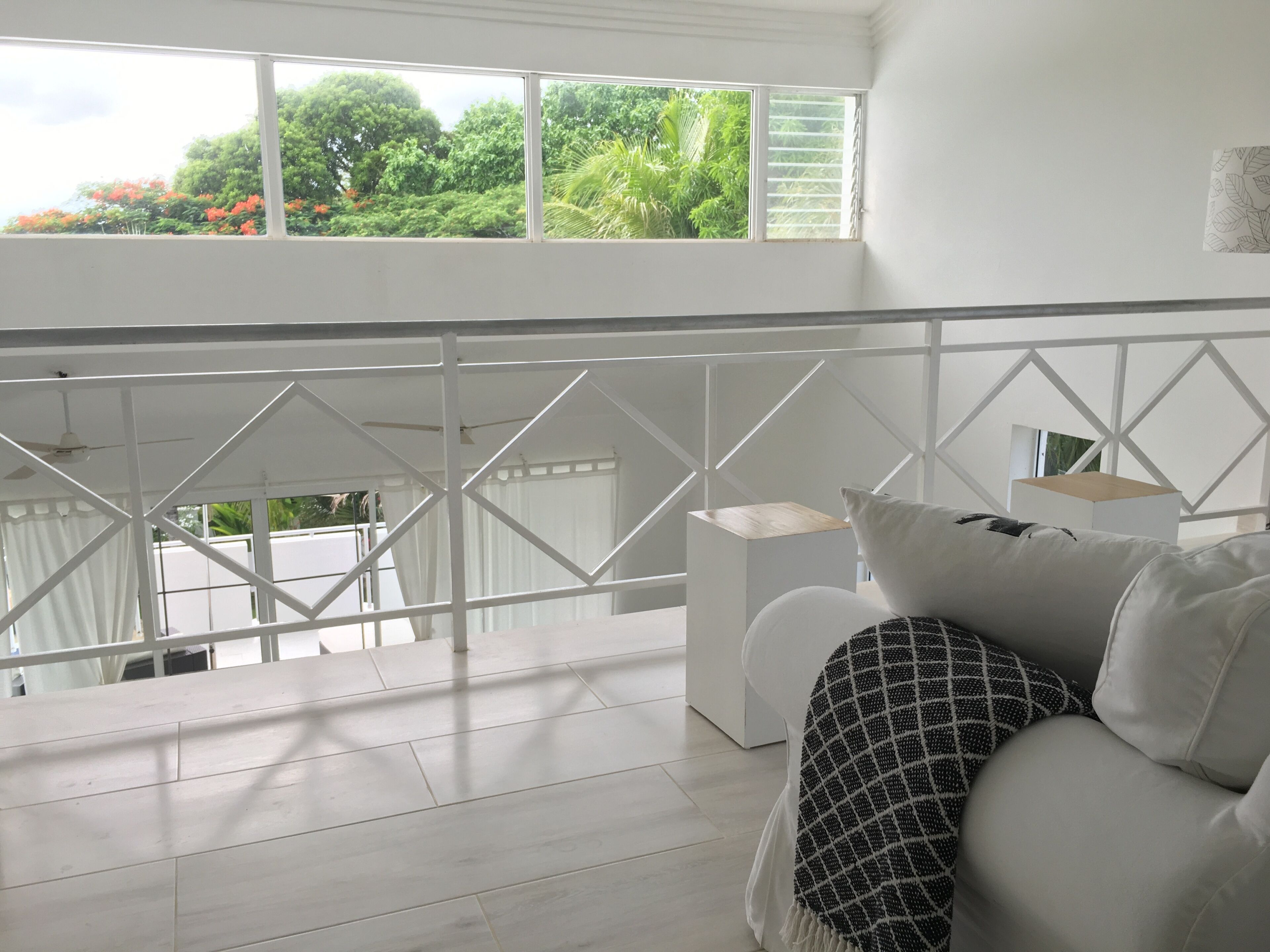 A bright room with a white balcony, a white sofa with patterned cushions, and a view of lush greenery outside.