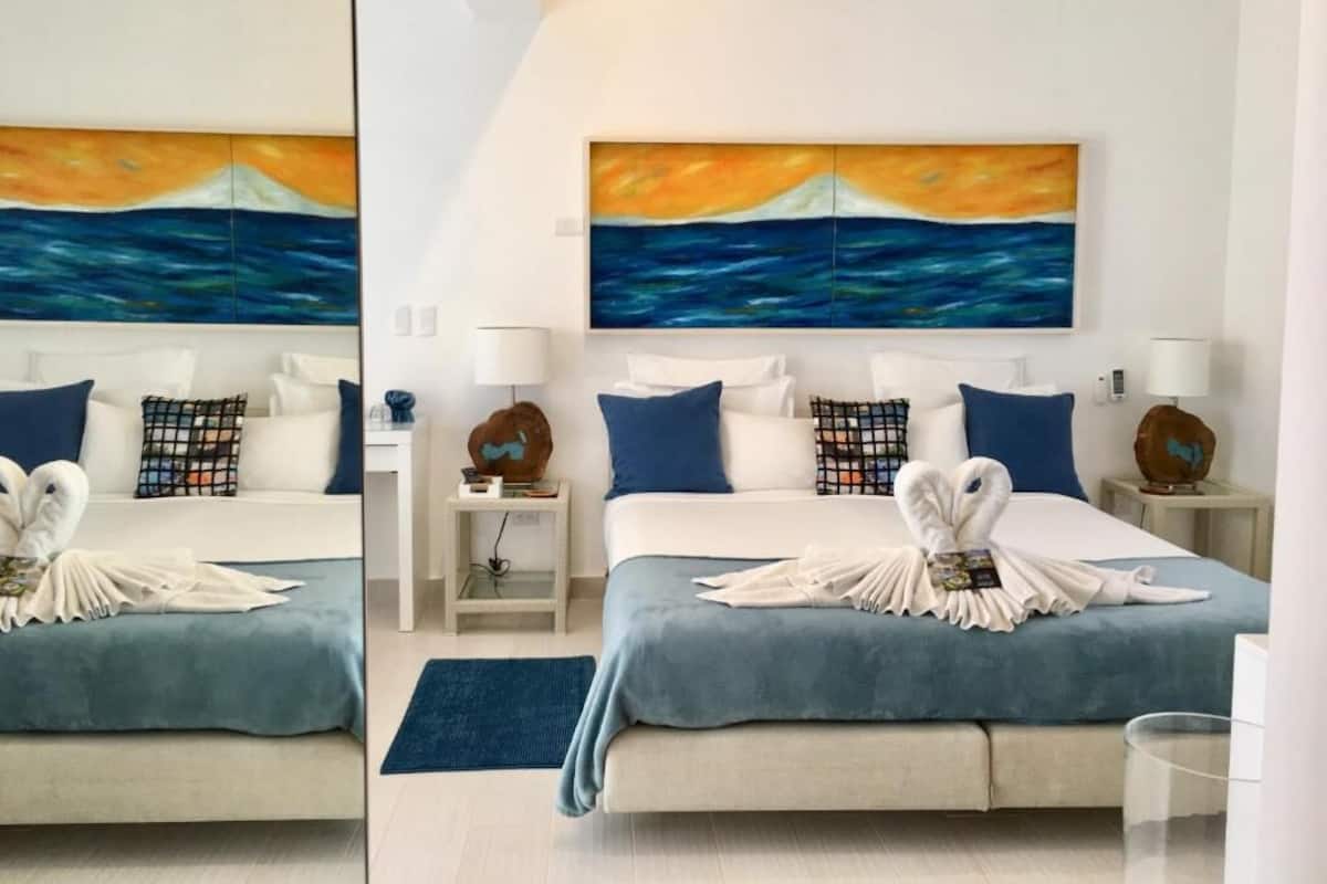 A hotel room with two beds, each with a blue bedspread, white pillows, and a matching blue pillow on each side. There are two paintings above the beds depicting a sunset over the ocean.