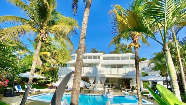 Zona de piscina con sillas de descanso, palmeras y un edificio de dos pisos al fondo.