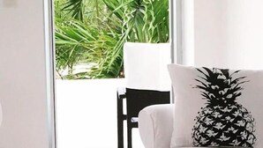 Una sala moderna con un sofá blanco y un cojín con motivos de piña, una silla negra y vistas a un follaje verde exuberante a través de un gran ventanal.
