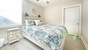 Une chambre à coucher comprenant un lit, un ventilateur de plafond, une fenêtre avec des stores, une table de chevet et une porte.