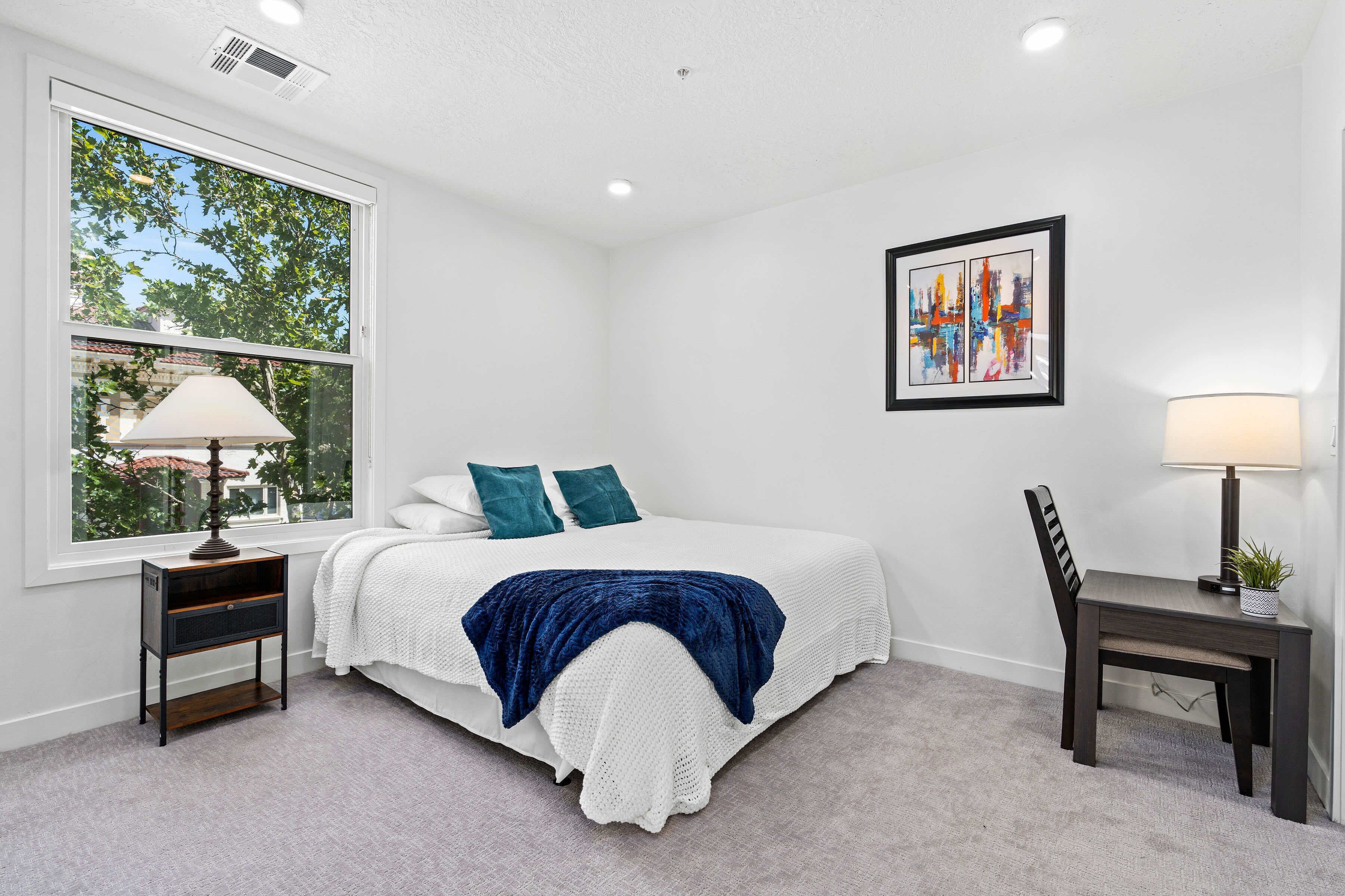 A bedroom with a bed, a desk, a chair, a window, and a painting on the wall.