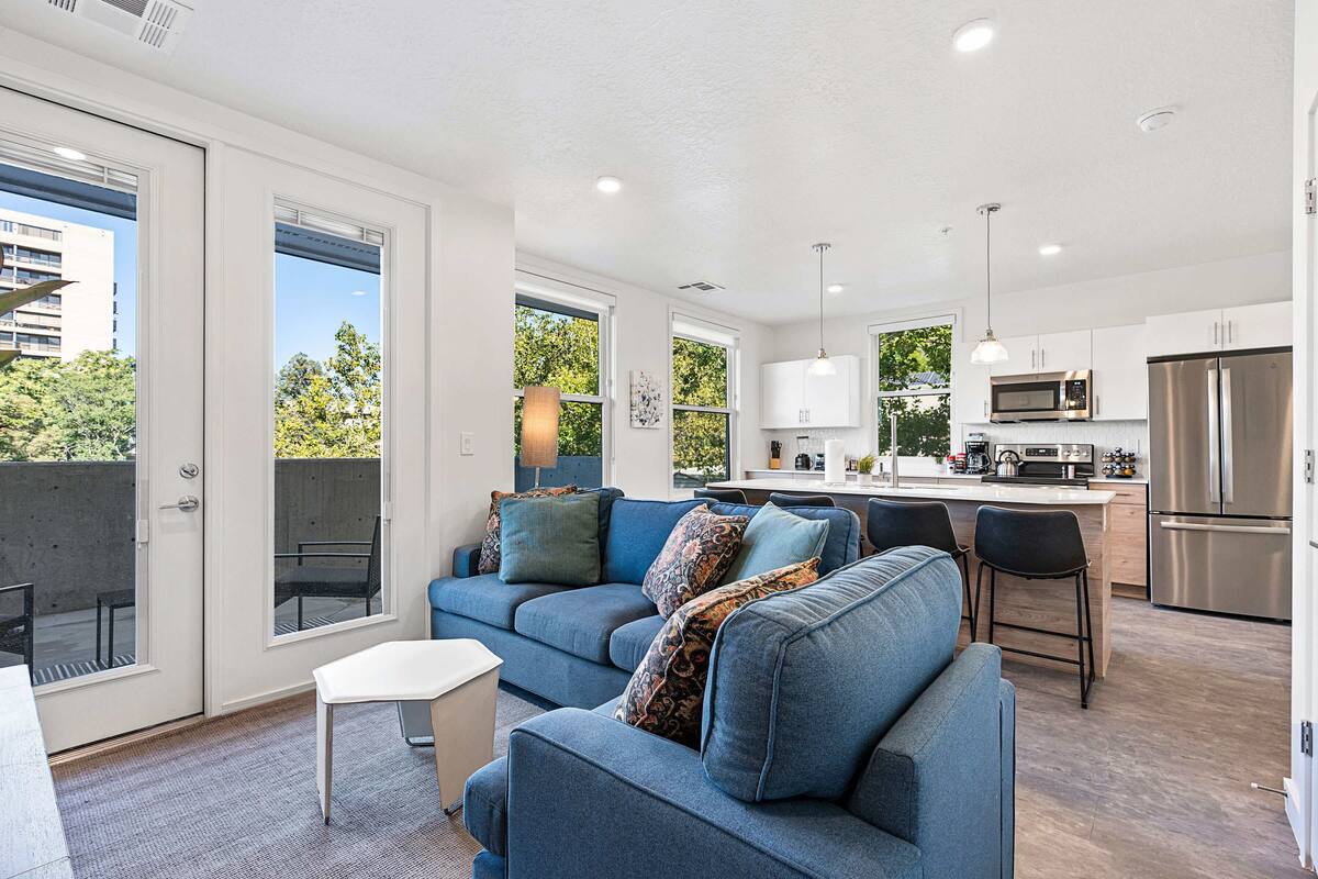 A modern living room with a blue sofa, a small white table, and a kitchen area with stainless steel appliances.
