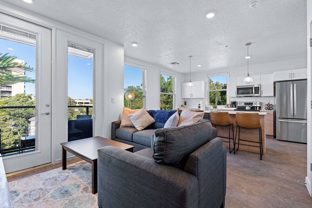 A modern living room with a grey sofa, a coffee table, and a dining area with bar stools. The kitchen features stainless steel appliances and white cabinetry.