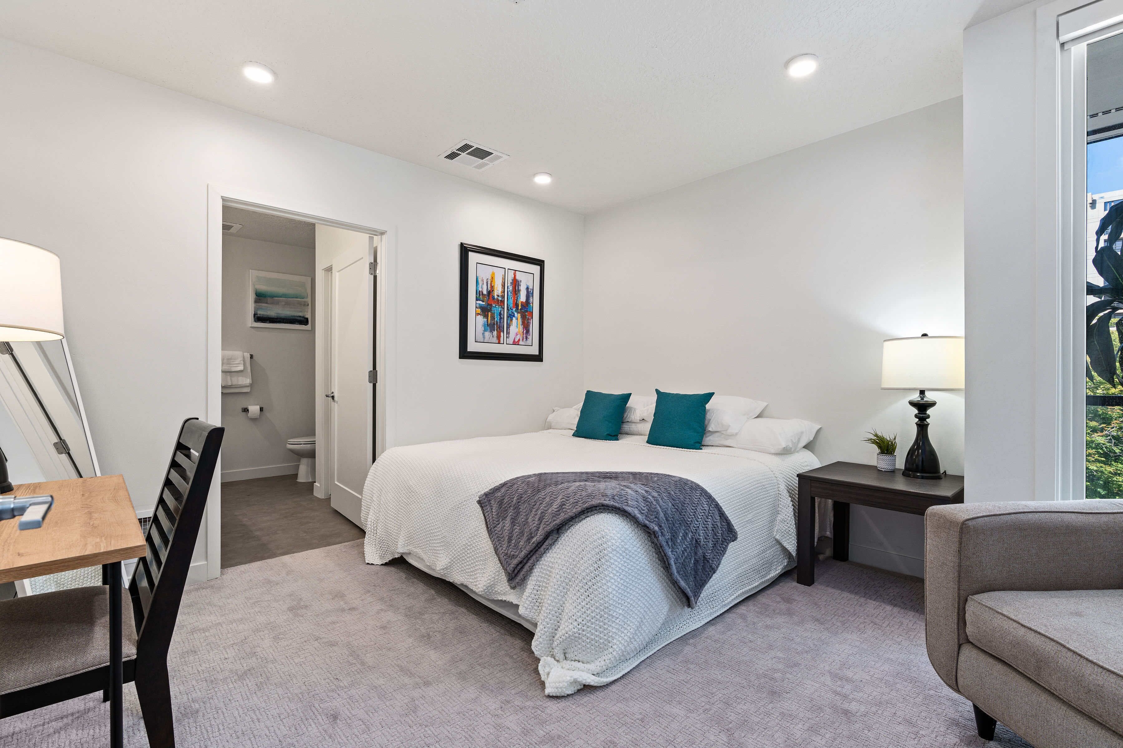 A bedroom with a bed, bedside table, lamp, and a view of the outdoors.