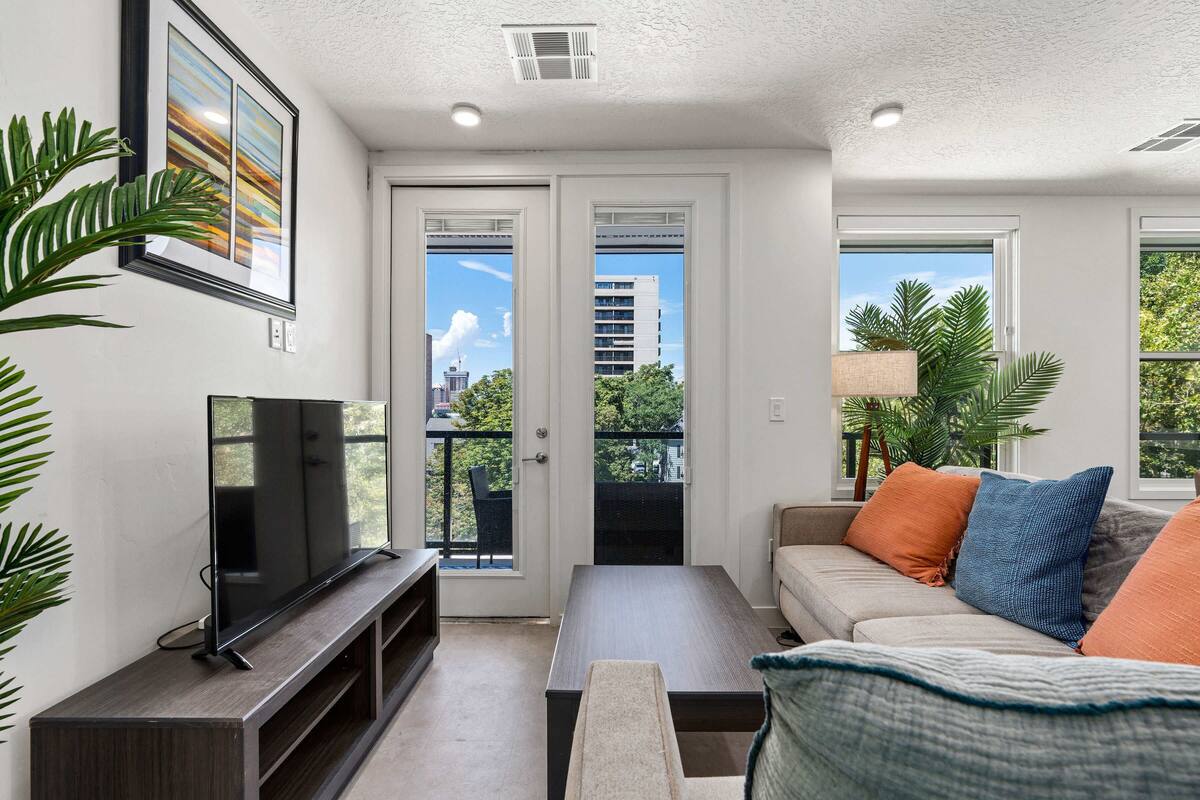A living room with a flat-screen TV, a sofa, a coffee table, and a door leading to a balcony with a view of buildings and trees.