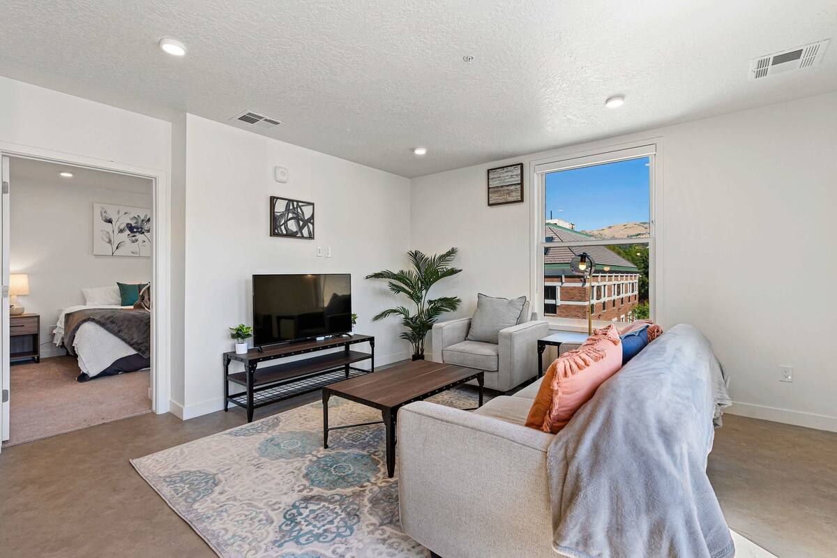 A modern living room with a flat-screen TV, a coffee table, and a sofa. There is a large window with a view of a building and hills.