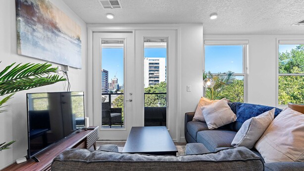 A modern living room with a flat-screen TV, a dark coffee table, and a grey sofa with multiple cushions. There is a large abstract painting on the wall and a potted plant to the left. - Luxury Apartment in Salt Lake City (Salt Lake City)