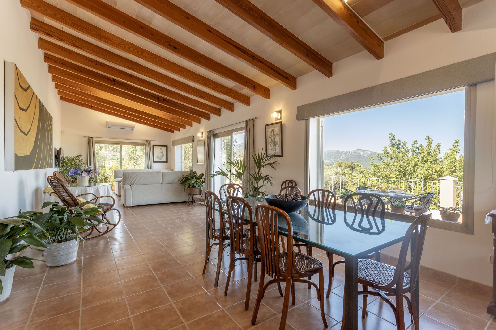 A dining area with a glass table and wooden chairs, a sofa, and a view of the outdoors.