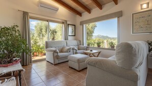 A spacious living room with a sofa, ottoman, and a small table. There are large windows with a view of trees and mountains.