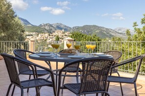 Une terrasse avec des chaises en osier et une table préparée pour le petit-déjeuner, d’où l’on peut admirer une ville nichée dans les montagnes.
