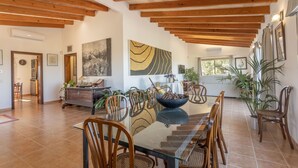 A spacious dining area with a long table and chairs, wooden beams on the ceiling, and potted plants.