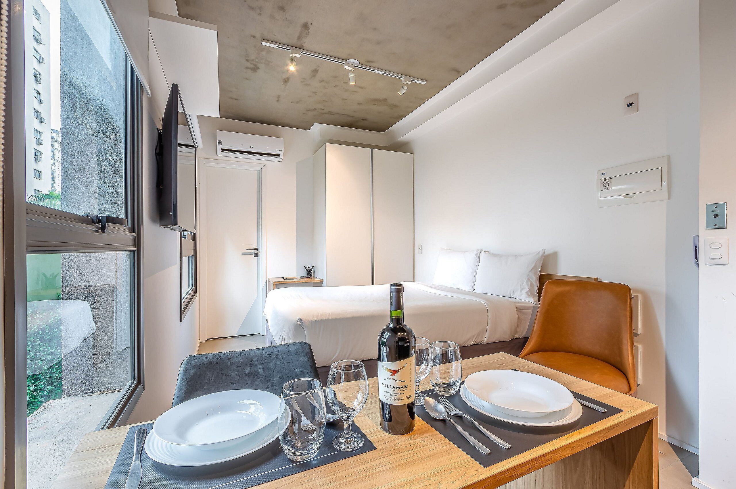 Habitación de hotel con una cama, una mesa de comedor con platos y vasos, una botella de vino y vistas a un edificio.