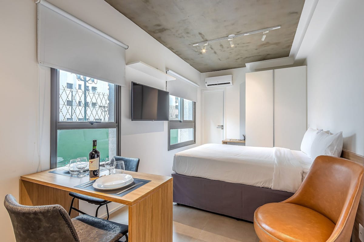 Una habitació d'hotel moderna amb un llit, una taula de treball amb una cadira, una televisió i una finestra que ofereix vistes a edificis.