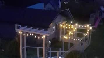 A small house with a porch and string lights, surrounded by a grassy area and a few trees.
