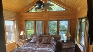Ein gemütliches Schlafzimmer in einer Holzhütte mit einem großen Bett, zwei Nachttischen, einem Deckenventilator und mehreren Fenstern mit Blick auf den umliegenden Wald.