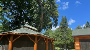Ein hölzerner Pavillon mit grüner Dachdecke, umgeben von Bäumen und einer Rasenfläche mit Sitzgelegenheiten.