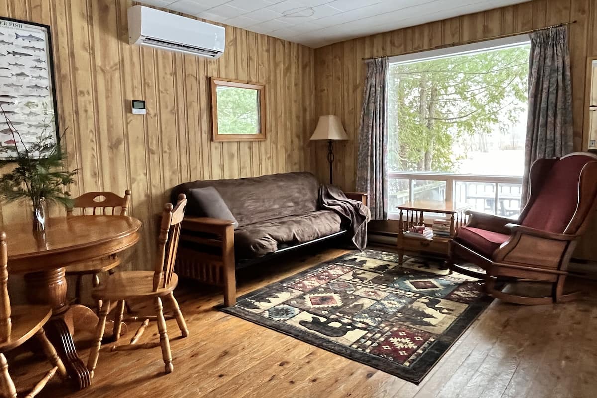 A cozy living room with a wooden floor, a brown leather sofa, a wooden dining table with chairs, a patterned rug, and a large window with curtains.