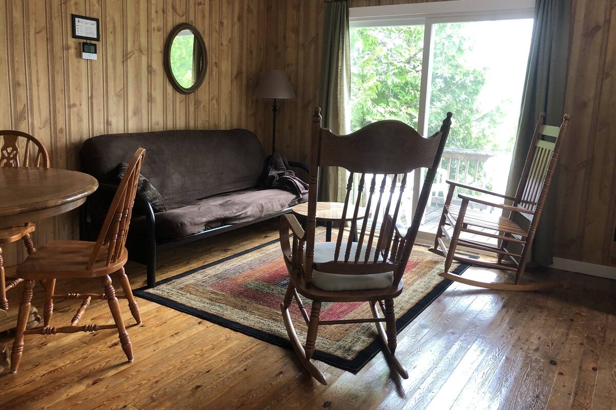 A cozy room with a wooden floor, a sofa, a rocking chair, a dining table with chairs, and a large window with curtains.