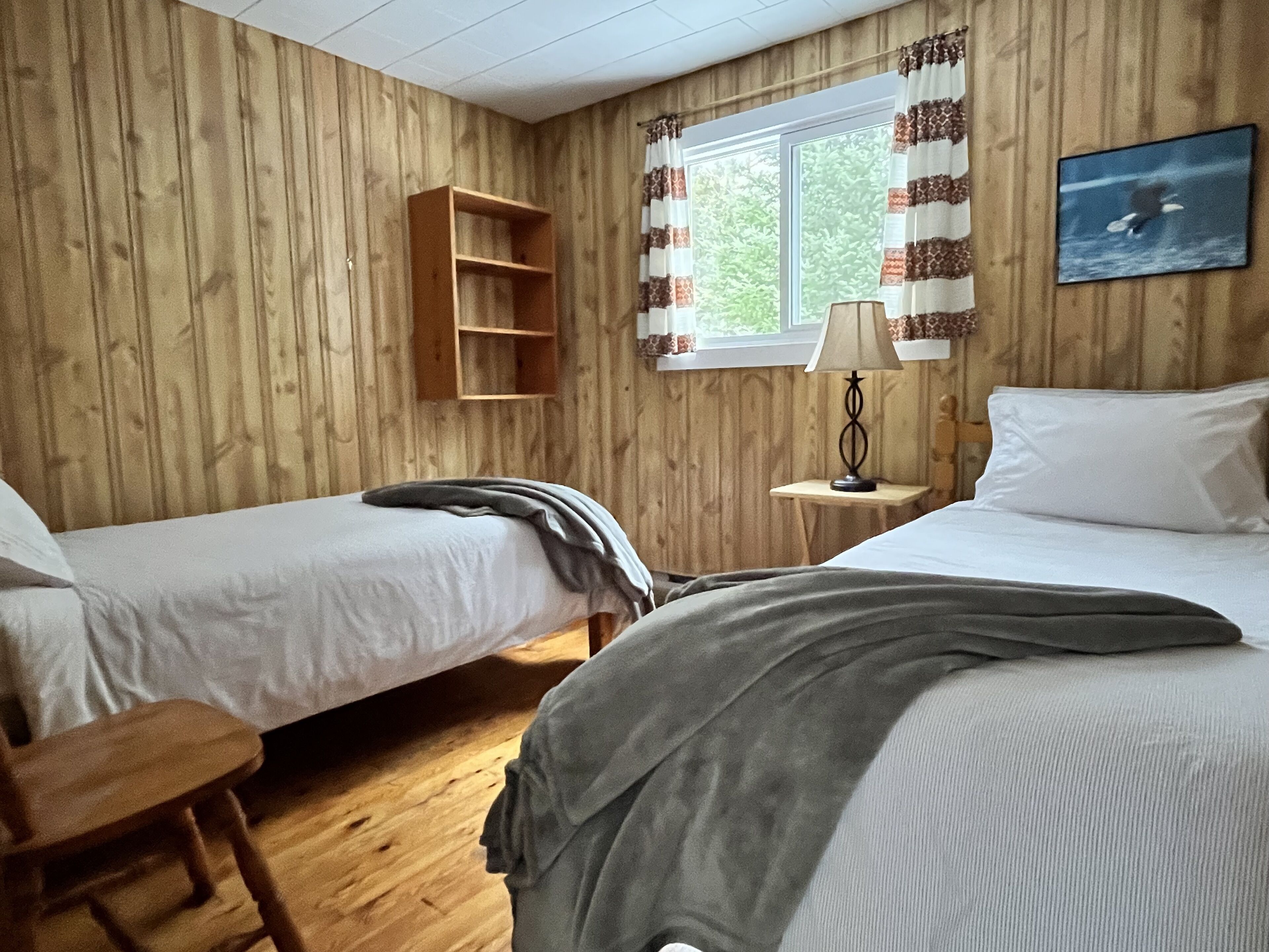 A room with two beds, a wooden shelf, a window with curtains, a lamp, and a picture on the wall.
