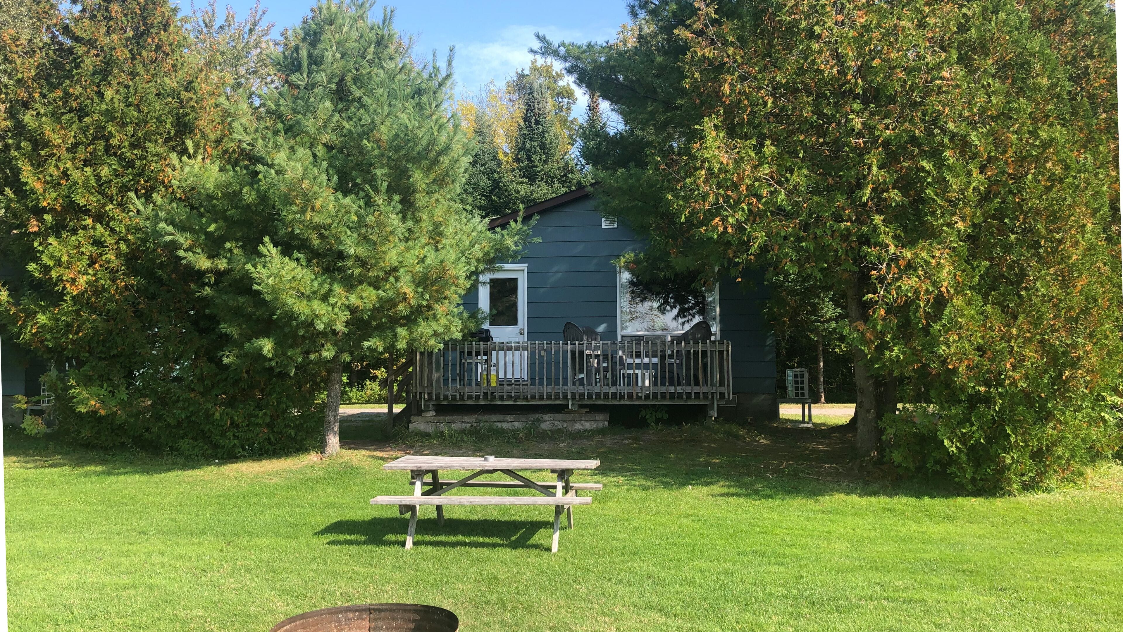 A cabin with a deck, surrounded by trees and a picnic table in a grassy area.