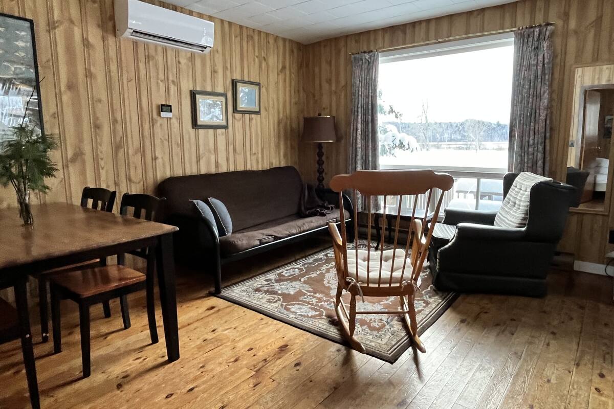 A cozy living room with wooden walls, a sofa, a rocking chair, a dining table, and a large window with curtains.