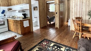 A compact kitchen with wooden cabinets, a sink, and a stove. There is a dining area with a wooden table and chairs. - Staying by the Lakeside Cottages (Plummer Additional)
